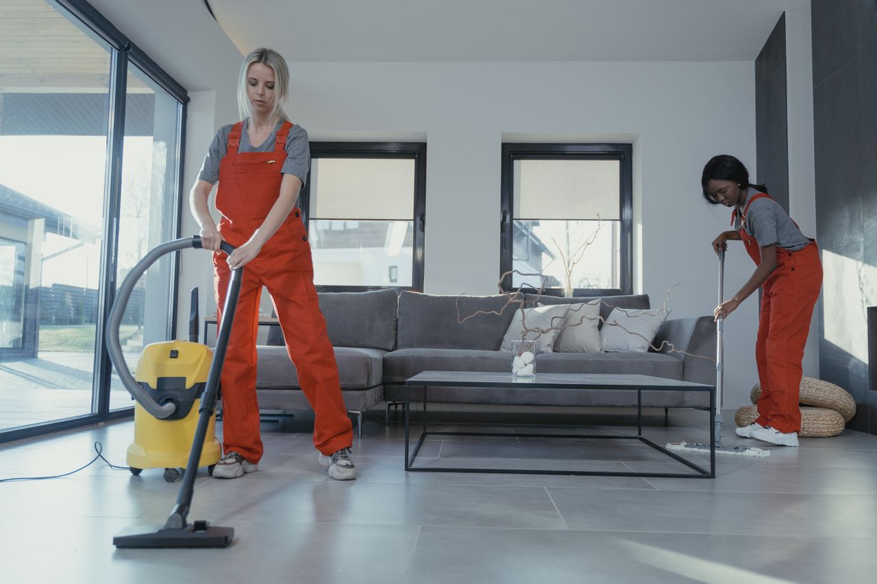 expertise-01 Two women in red overalls vacuuming and sweeping a modern living room.
